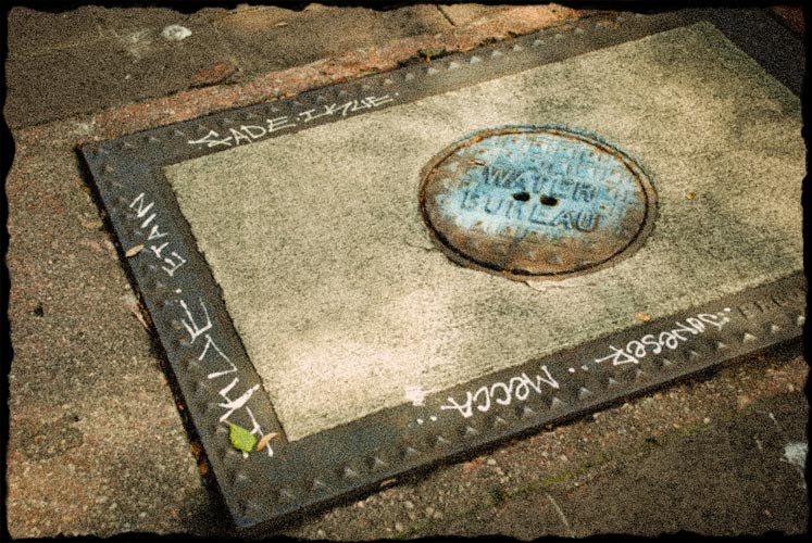 Street water meter cover with graffiti all around the edges.